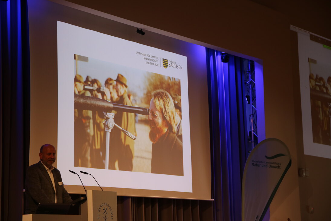 Dr. Rolf Tenholtern steht auf der Bühne und spricht. Im Hintergrund sind Bilder von Helmut Ballmann zu sehen.