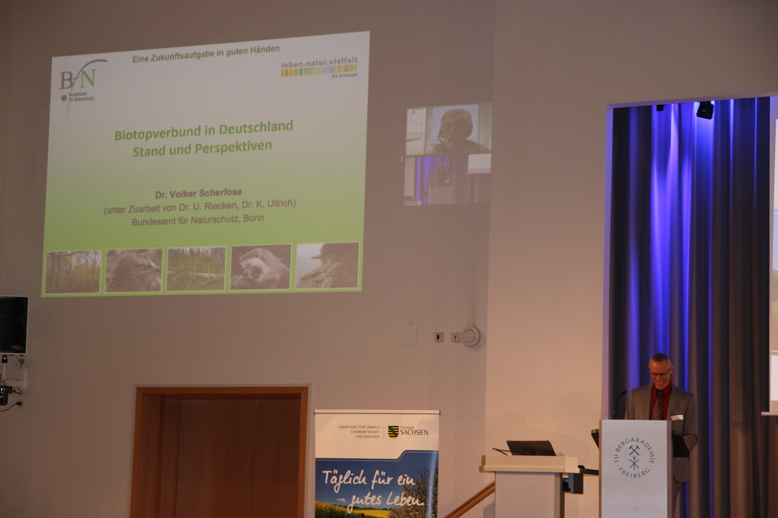 Auf der Wand neben der Bühne ist die Präsentation von Dr. Volker Scherfose zu sehen, er ist in einem kleinen Bild eingeblendet. Dr. Scherfose referierte zum Thema »Biotopverbund in Deutschland – Stand und Perspektiven«.